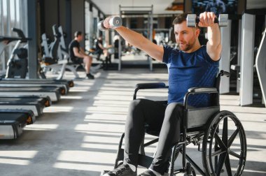 Spor salonunda tekerlekli sandalyede halterle egzersiz yapan engelli bir adam rehabilitasyon görüyor.