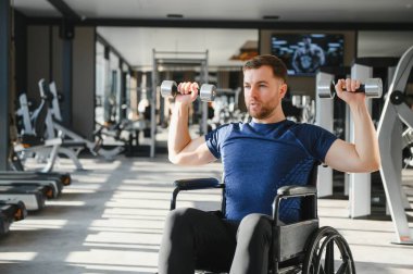 Özürlü bir adam, sporcu spor salonunda rehabilitasyonu sırasında tekerlekli sandalyede otururken kollarını kaldırarak egzersiz yapıyor.