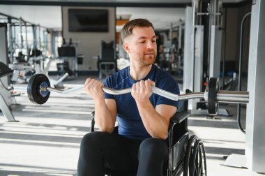 Tekerlekli sandalyedeki beyaz adam, parlak bir spor salonunda ağırlık kaldırıyor, rehabilitasyon ya da fizyoterapi sırasında pazılarını çalıştırıyor.