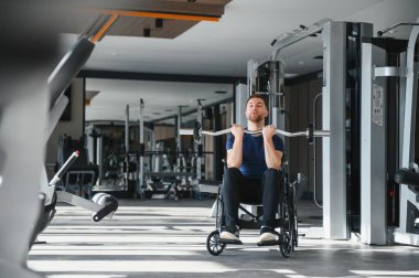 Adam spor salonunda tekerlekli sandalyede ağırlık kaldırıyor, egzersiz yaparak güç, esneklik ve rehabilitasyon gösteriyor.