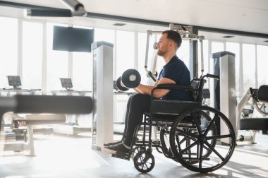 Tekerlekli sandalyede otururken dambıllarla egzersiz yapan, bir spor salonunda fiziksel aktivite yoluyla rehabilitasyonu teşvik eden kişi.