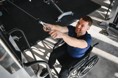 Tekerlekli sandalyedeki bir adam spor salonunda dayanıklılık eğitimi alıyor, rehabilitasyona ve özel ekipmanlarla spor yapmaya odaklanıyor.