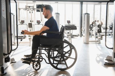 Tekerlekli sandalyedeki adam spor aletlerini rehabilitasyon ve spor eğitimi için iyi aydınlatılmış bir spor merkezinde aktif olarak kullanıyor.