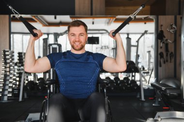 Modern bir spor salonunda ağırlık kaldıran tekerlekli sandalyedeki gülümseyen adam azim ve güç gösterisi yapıyor.