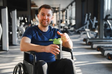 Tekerlekli sandalyedeki adam sallama şişesini spor salonunda tutuyor. Spor rehabilitasyonunu ve engelliler için aktif yaşam tarzını simgeliyor.