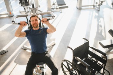 Engelli bir adam, modern spor salonunda ağırlık egzersizi yapıyor. Dayanıklılık, dayanıklılık ve kendini tekerlekli sandalyesiyle spor yapmaya adamış.
