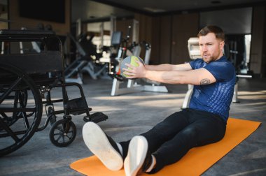 Özürlü bir adam jimnastik paspası üzerinde tıp topuyla egzersiz yapıyor rehabilitasyona ve aktif yaşam tarzına bağlılığını gösteriyor.