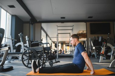 Spor salonundaki paspasın üzerinde egzersiz yapan engelli bir adam, tekerlekli sandalyesi yanında, azim ve güç gösterisi yapıyor.