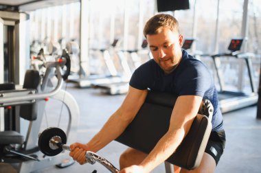 Odaklanmış erkek sporcu, modern bir spor salonundaki bir makinenin üzerinde biceps kıvrımları ile fiziksel rehabilitasyonu ve iyileşmesi üzerinde çalışıyor.