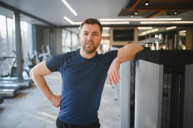 Spor salonunda antrenman yaptıktan sonra dinlenen sporcu, spor makinesine yaslanıyor, özgüven ve kararlılık gösteriyor.