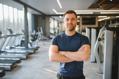 Aydınlık bir spor salonunda kolları çapraz duran bir fitness hocasının portresi, fitness endüstrisinde profesyonellik ve uzmanlık gösteriyor.