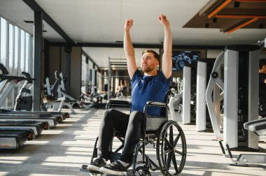 Tekerlekli sandalyede egzersiz yapan, modern spor salonunda rehabilitasyon sırasında kollarını kaldıran bir adam.