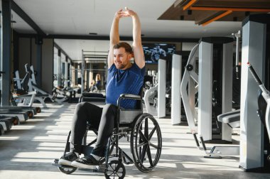 Modern bir spor salonunda tekerlekli sandalyede oturan engelli, egzersiz yapan ve kollarını esneten biri.