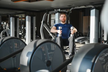 Tekerlekli sandalyedeki bir adam spor salonunda dambıllarıyla egzersiz yapıyor, rehabilitasyon ve zindeliğe odaklanıyor. Teçhizat ve ağırlık arkaplanda görünür