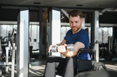 Dambıllarla egzersiz yapan, spor salonunda tekerlekli sandalyede oturan, rehabilitasyon gören ve fiziksel durumunu iyileştiren bir adam.