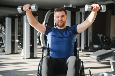 Tekerlekli sandalyedeki gülümseyen adam spor salonundaki rehabilitasyon sırasında ağırlık kaldırıyor. Güç, azim ve uyum sağlama yeteneği gösteriyor.