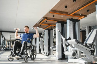 Modern spor salonunda dambıllarla egzersiz yapan engelli bir adam spor yaparak kapsayıcılığını ve rehabilitasyonunu gösteriyor.