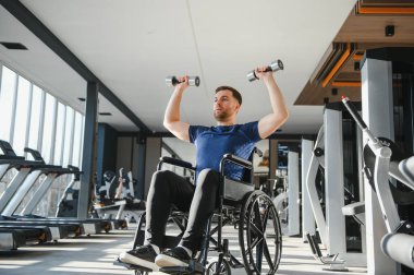 Tekerlekli sandalyedeki engelli adam dambıl kaldırıyor, modern spor salonunda egzersiz yapıyor, rehabilitasyon, spor ve aktif yaşam tarzına bağlılığını gösteriyor.