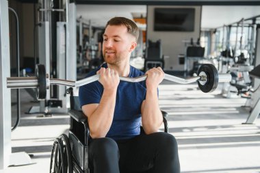 Özürlü bir adam, tekerlekli sandalyede ağırlık kaldıran bir sporcu, parlak, modern bir spor salonunda rehabilitasyon sırasında güç ve direnç gösteriyor.