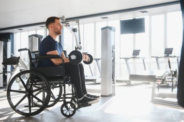 Tekerlekli sandalyedeki kişi, halterlerle egzersiz yapıyor, sporu kapsayacağını ve formda kalacağını gösteriyor.