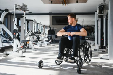 Engelli genç sporcu tekerlekli sandalyede oturuyor ve modern spor salonunda halter antrenmanına hazırlanıyor.