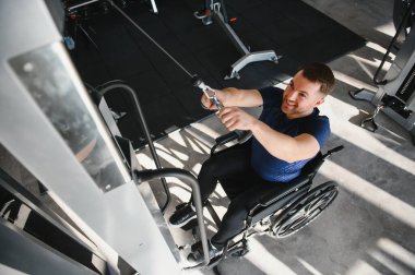 Tekerlekli sandalyedeki gülümseyen adam kollarını spor salonunda çekiş makinesiyle çalıştırıyor. Fitness ve rehabilitasyona bağlılığını gösteriyor.