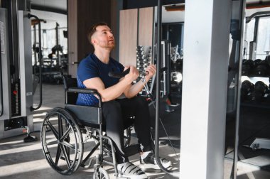 Tekerlekli sandalyedeki kararlı erkek sporcu kollarını ağırlık makinesiyle çalıştırıyor, ulaşılabilir spor salonunda dayanıklılık ve güç gösteriyor.