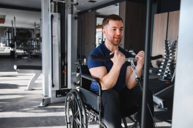 Tekerlekli sandalyedeki adam kollarını spor salonunda ağırlık makinesiyle çalıştırıyor. Engelliler için rehabilitasyon ve uygunluk gösterisi yapıyor.