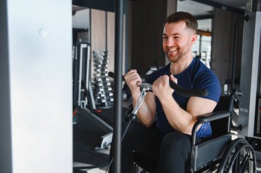 Tekerlekli sandalyedeki gülümseyen adam ağırlık makinesi kullanıyor, modern spor salonunda pazılarını çalıştırıyor, rehabilitasyon ya da spor yaparken azim ve güç gösteriyor.