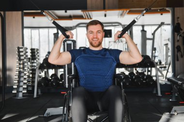 Tekerlekli sandalyedeki gülümseyen adam rehabilitasyon sırasında ağırlık kaldırıyor, modern bir spor salonunda azim ve güç gösterisi yapıyor.