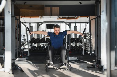 Tekerlekli sandalyedeki adam kablo ağırlıklarıyla egzersiz yapıyor. Erişilebilir spor salonundaki rehabilitasyon eğitiminde güç ve direnç gösteriyor.
