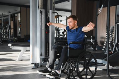 Tekerlekli sandalyedeki adam üst vücut egzersizi için kablo kullanıyor. Modern spor salonunda rehabilitasyon sırasında güç ve direnç gösteriyor.