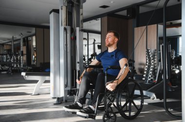 Tekerlekli sandalyedeki adam spor salonunda kablo ağırlığıyla egzersiz yapıyor. Rehabilitasyon ve zindeliğe katkı ve azim gösteriyor.