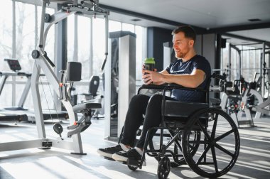 Tekerlekli sandalyedeki gülen adam spor salonunda egzersiz yaptıktan sonra protein içeceği içiyor. Kararlılık ve sağlıklı yaşam tarzı gösteriyor.
