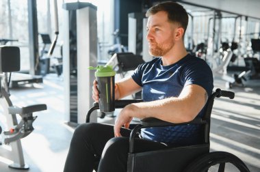 Tekerlekli sandalyedeki adam, modern spor salonundaki rehabilitasyon egzersizlerinden sonra dinlenirken bir protein karıştırıcı tutuyor.