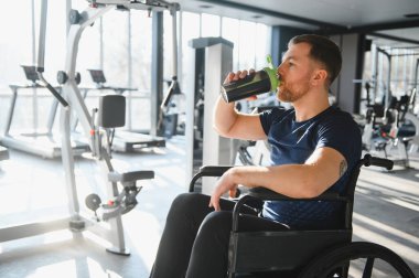 Engelli erkek sporcu protein içeceği içiyor, modern spor salonundaki yoğun eğitimden sonra tekerlekli sandalyede dinleniyor.