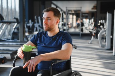 Engelli erkek sporcu, modern bir spor salonunda egzersiz yaptıktan sonra elinde su şişesiyle tekerlekli sandalyesinde mola veriyor.