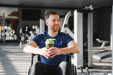 Modern spor salonundaki rehabilitasyon sırasında elinde protein karıştırıcı ve gülümseyen tekerlekli sandalyedeki mutlu adam.