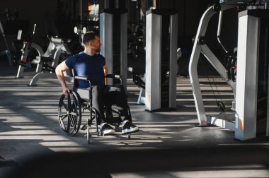 Modern bir spor salonunda tekerlekli sandalyede egzersiz yapan engelli bir adam, spor ve rehabilitasyon sürecinde direnç ve kararlılık gösteriyor.