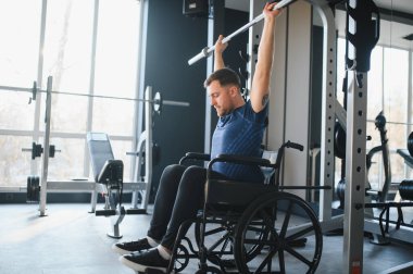 Erkek sporcu tekerlekli sandalyede otururken ağırlık kaldırıyor, modern spor salonunda egzersiz yapıyor ve rehabilitasyon için antrenman yapıyor.