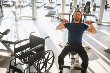 Adam ağırlık makinesi kullanıyor, tekerlekli sandalyeyle spor salonunda egzersiz yapıyor, spor rehabilitasyonu ve kapsamı gösteriyor.
