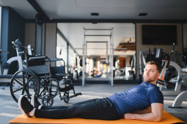 Özürlü bir adam spor salonundaki paspasın üzerinde, tekerlekli sandalyenin yanında, rehabilitasyon sırasında azim ve güç gösteriyor.