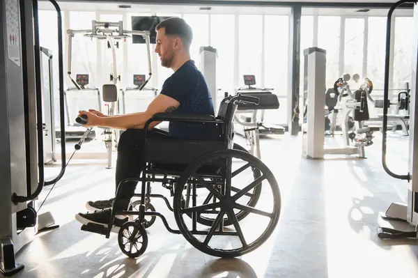 Tekerlekli sandalyedeki adam spor aletlerini rehabilitasyon ve spor eğitimi için iyi aydınlatılmış bir spor merkezinde aktif olarak kullanıyor.