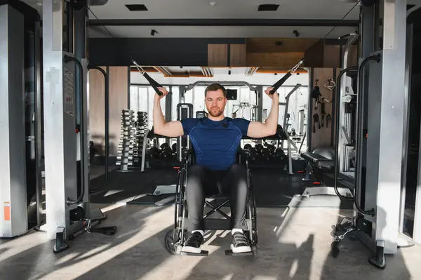 Tekerlekli sandalyedeki kablo makinesini kullanan erkek sporcu spor salonundaki rehabilitasyon sırasında ağırlık kaldırıyor ve egzersiz yapıyor.