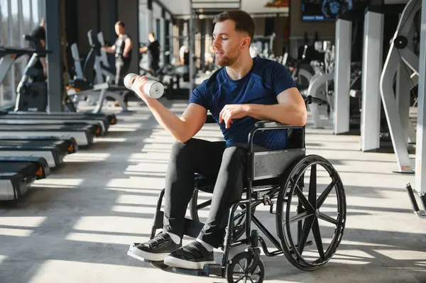 Spor salonunda engelli bir kişi, güç eğitimi için dambıl kullanıyor, spor rehabilitasyonunu ve kendini adamışlığı vurguluyor.