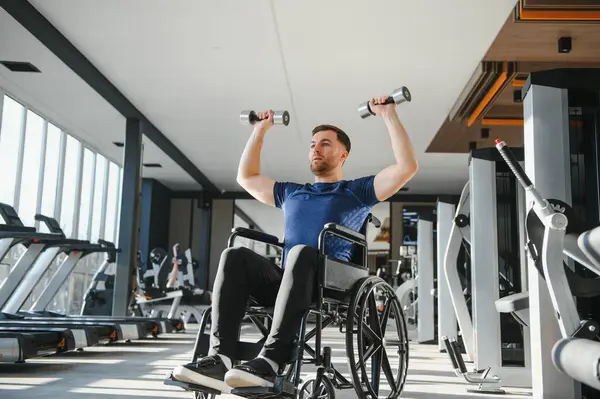 Tekerlekli sandalyedeki engelli adam dambıl kaldırıyor, modern spor salonunda egzersiz yapıyor, rehabilitasyon, spor ve aktif yaşam tarzına bağlılığını gösteriyor.