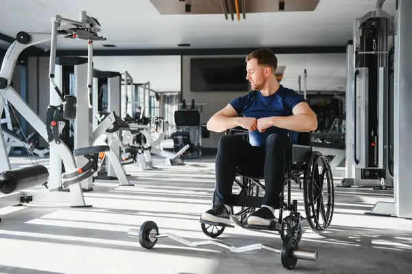 Engelli genç sporcu tekerlekli sandalyede oturuyor ve modern spor salonunda halter antrenmanına hazırlanıyor.