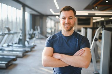 Çapraz kollarla gülümseyen kendine güvenen fitness eğitmeni modern bir spor salonunda profesyonellik ve uzmanlık gösteriyor.