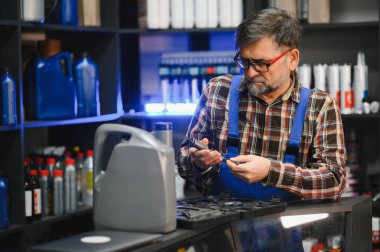 Kıdemli satıcı mavi tulumlar giyip bir oto yedek parça mağazasında küçük bir parça tutuyor ve nesneye odaklanıyor.