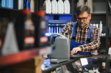 Kıdemli satıcı mavi tulum giyer ve oto yedek parça dükkanında kasayı kullanır. Motor yağı ve tezgahta aletler vardır.